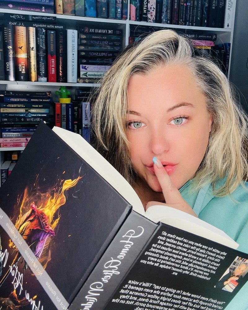 J L Yock holding a finger to her lips behind an open copy of Home After Midnight