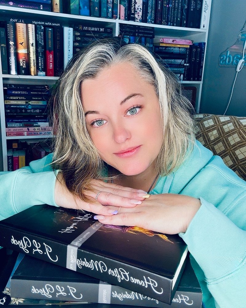 J L Yock resting her hands on a stack of her Home After Midnight hardbacks in front of her bookshelves