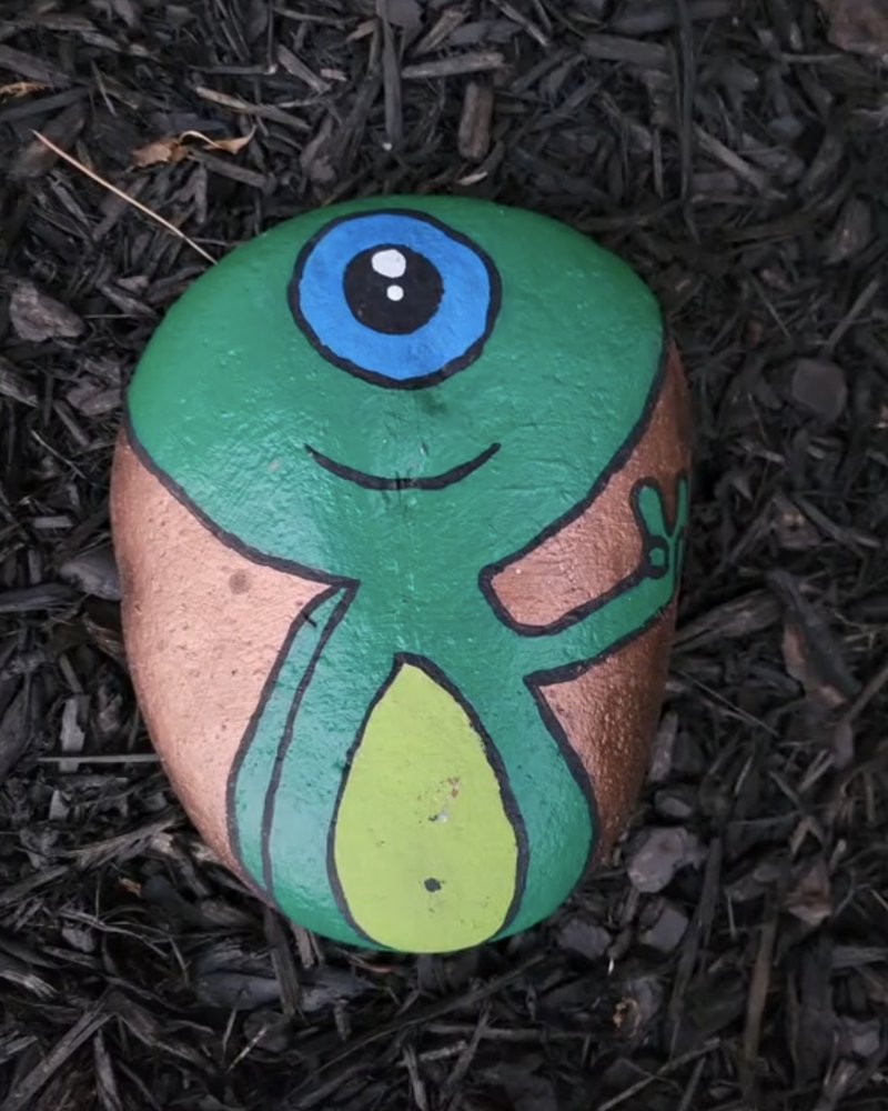 Big handpainted art rock: a smiling green one-eyed creature giving a peace sign